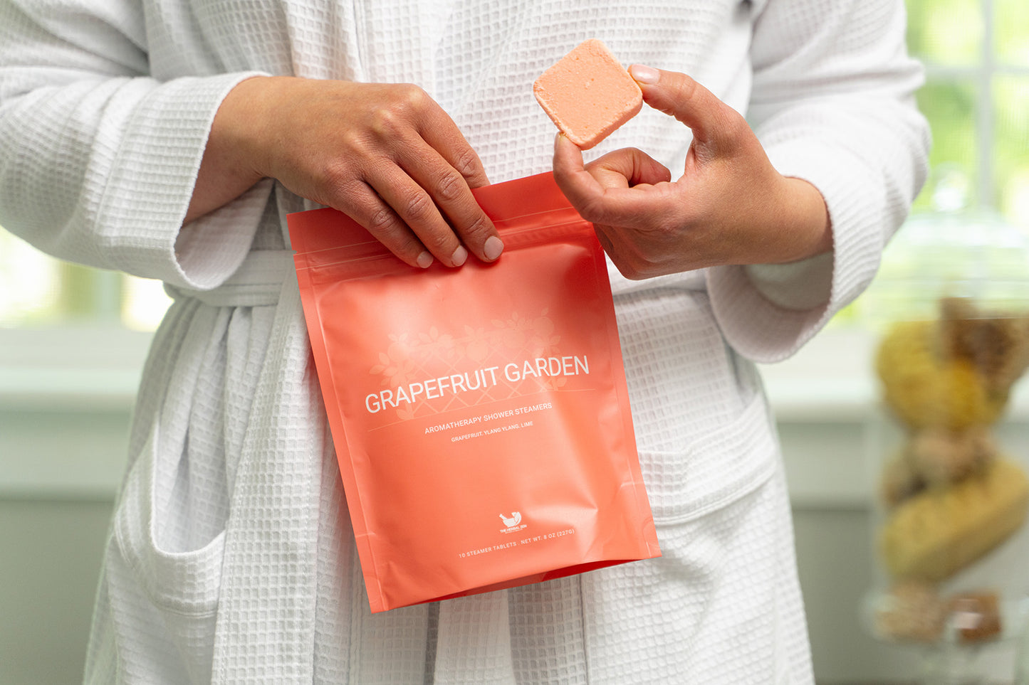 Person holding a package of Grapefruit Garden shower steamer by The Herbal Zen in their right hand and a single shower steamer in their left hand.